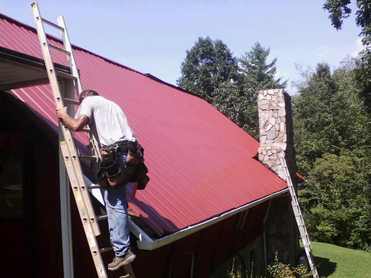 Roofer on a ladder beside a red metal roof and stone chimney — Soffit & Fascia Repair by Roofing Brothers in Kensett